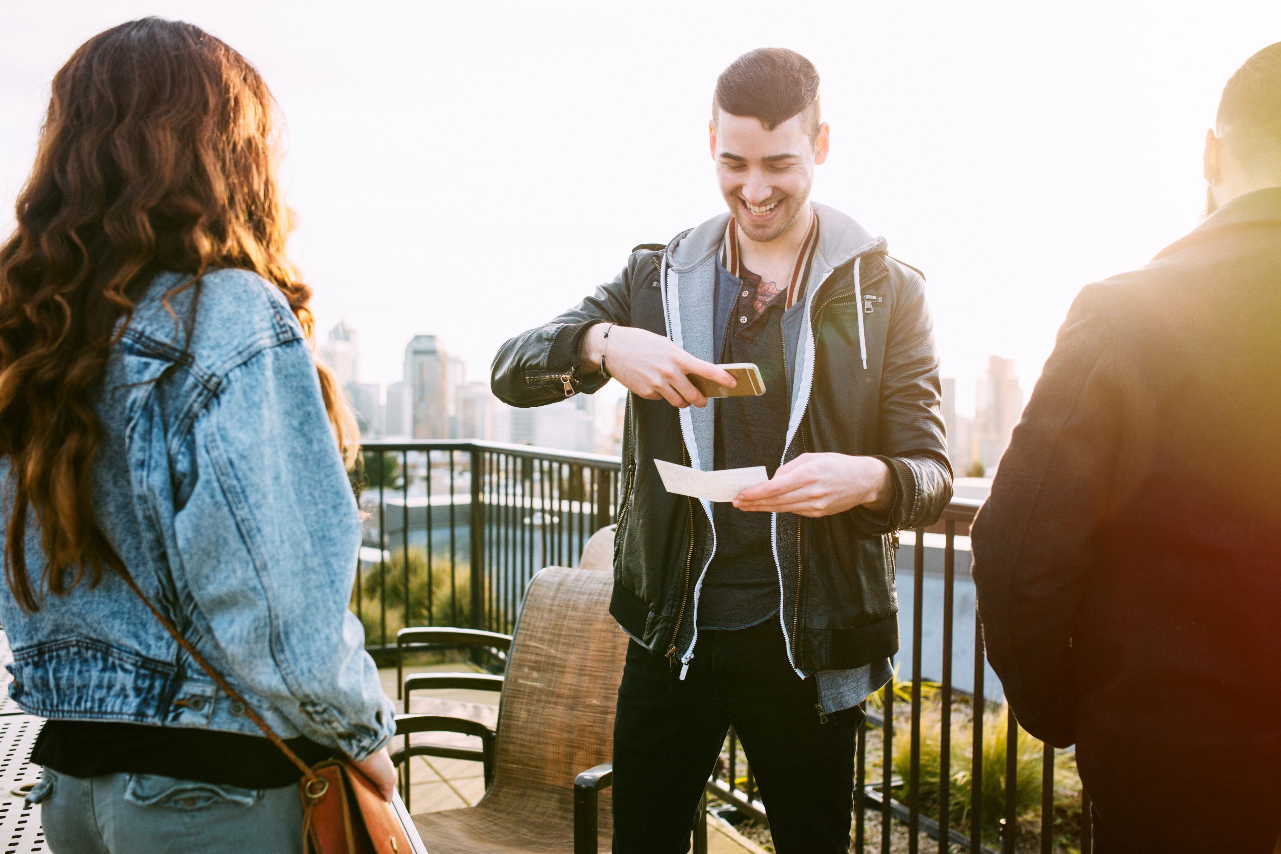 A man on a Seattle roof top taking a picture of a check with his smart phone for a Remote Deposit Capture.  He smiles, wearing modern stylish clothing, his friends waiting for him to finish the transaction.  Seattle cityscape on the skyline in background.